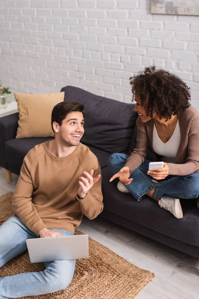 Positive multiethnic couple with devices gesturing in living room — Stock Photo