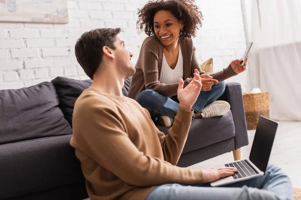 Freelancer with laptop talking to african american girlfriend with cellphone at home — Stock Photo