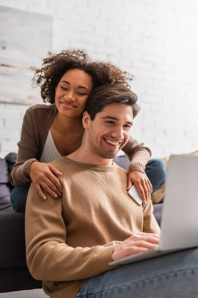 Mujer afroamericana joven con novio abrazador de teléfono celular usando un ordenador portátil borroso en casa - foto de stock