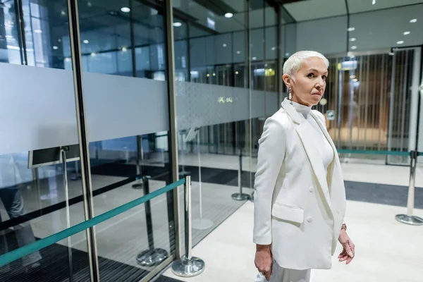 Senior business lady en traje blanco de pie en hotel moderno cerca de la entrada - foto de stock