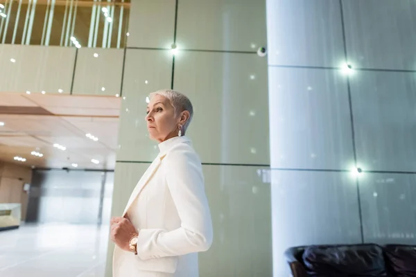 Vista lateral da mulher de negócios sênior loira em blazer branco no lobby do hotel moderno — Fotografia de Stock
