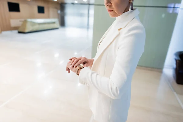 Vista recortada de la rica mujer de negocios comprobando el tiempo en el reloj de pulsera de oro en el vestíbulo del hotel - foto de stock