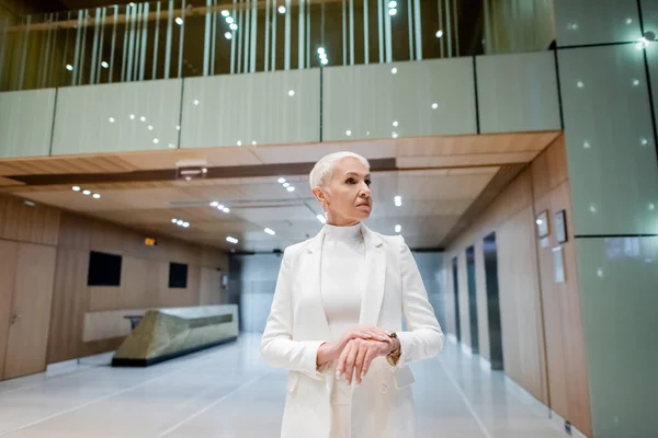 Mature businesswoman in white trendy clothes looking away in lobby of modern hotel — Stock Photo