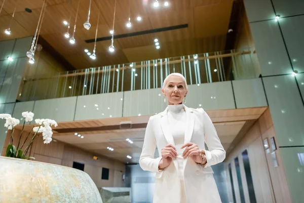 Low angle view of senior businesswoman in white stylish blazer looking at camera in luxury hotel — Stock Photo