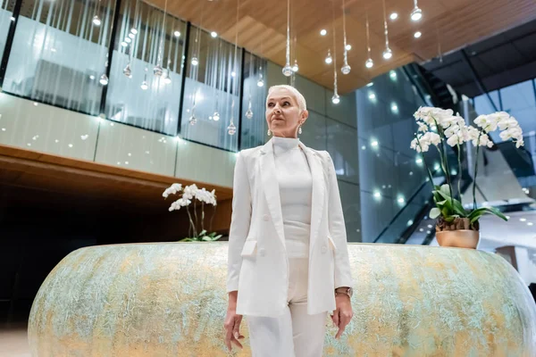 Blonde senior businesswoman in white clothes standing near orchids in luxury hotel — Stock Photo