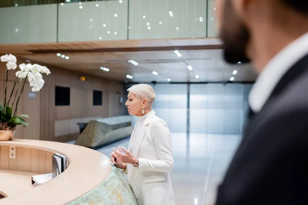 Trendy senior businesswoman near hotel reception and bi-racial bodyguard on blurred foreground — Stock Photo