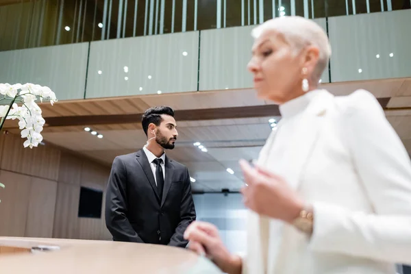 Двурасовый телохранитель смотрит в сторону, пока размытая деловая леди регистрируется на стойке регистрации отеля — стоковое фото