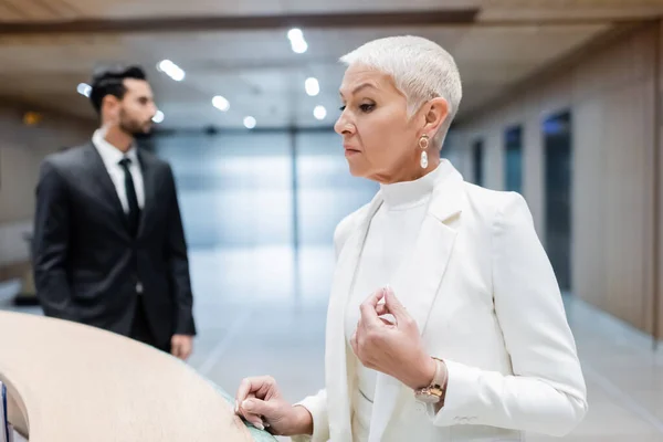 Senior and wealthy businesswoman checking in on hotel reception near blurred bi-racial bodyguard — Stock Photo