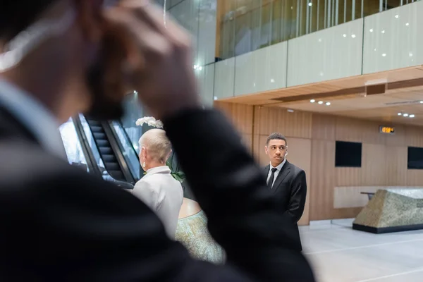 Guardaespaldas afroamericano protegiendo a mujer de negocios senior en hotel cerca de hombre de seguridad bi-racial en primer plano borroso - foto de stock