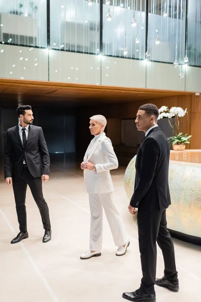 Vue pleine longueur de femme d'affaires mature et à la mode marchant près de gardes du corps interracial privés dans le hall de l'hôtel — Photo de stock