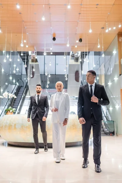 Full length view of stylish senior business lady walking near private interracial bodyguards in luxury hotel — Stock Photo