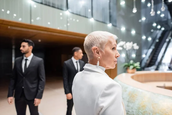 Mature and stylish businesswoman looking away near interracial bodyguards in modern hotel — Stock Photo