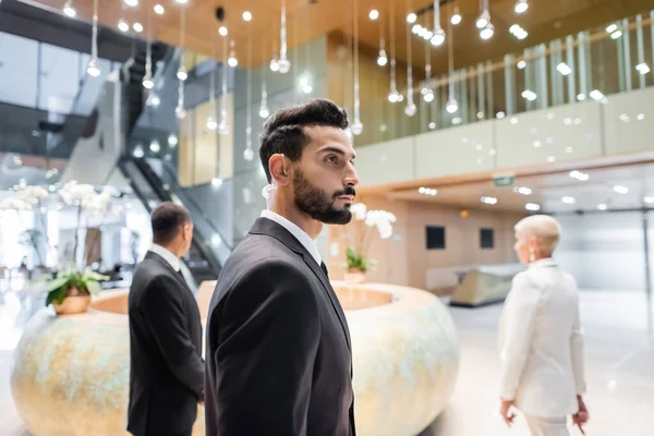 Mixed race bodyguard in security earpiece looking away while escorting senior businesswoman in hotel — Stock Photo