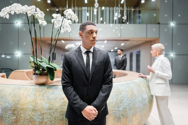 Hombre de seguridad afroamericano mirando hacia otro lado cerca borrosa mujer de negocios en la recepción del hotel - foto de stock