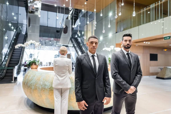 Interracial bodyguards looking at camera near senior business lady checking in at hotel reception — Stock Photo