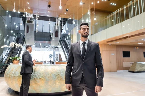 Hombre de seguridad bi-racial en traje de pie en el vestíbulo del hotel cerca de la guardia afroamericana en la recepción - foto de stock