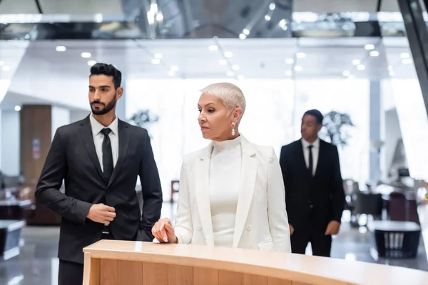 Adinerada dama de negocios esperando en la recepción del hotel cerca de guardaespaldas interracial - foto de stock