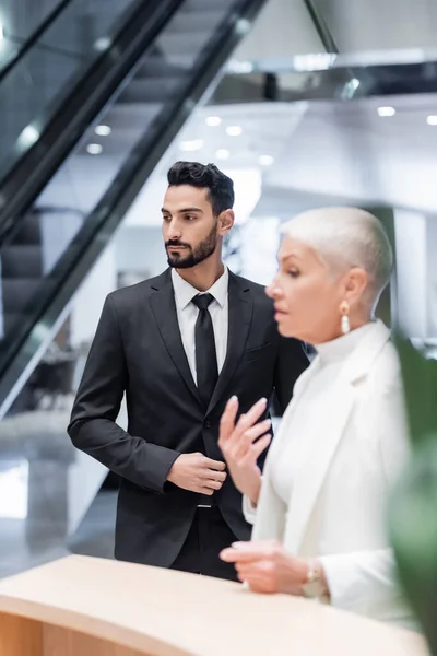 Joven bi-racial seguridad hombre en formal desgaste cerca borrosa senior businesswoman en hotel - foto de stock