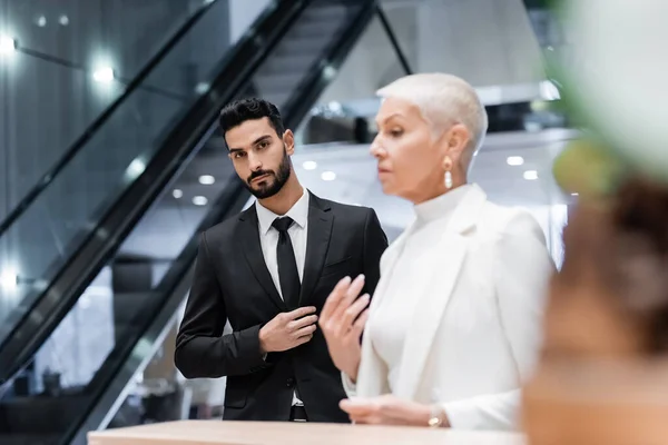 Giovane bi-razziale uomo di sicurezza guardando la fotocamera vicino offuscata signora d'affari in attesa alla reception dell'hotel — Foto stock