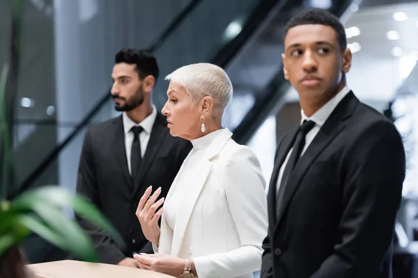 Stylish senior business lady near hotel reception and private multiethnic bodyguards — Stock Photo