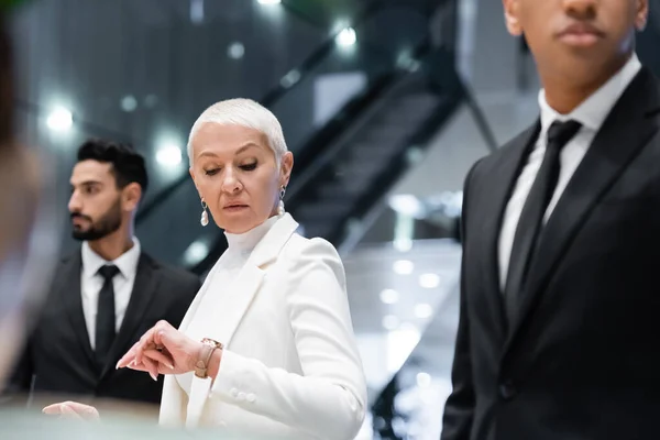 Reife Geschäftsfrau überprüft Zeit auf Armbanduhr in der Nähe von interrassischen Leibwächtern im Hotel — Stockfoto