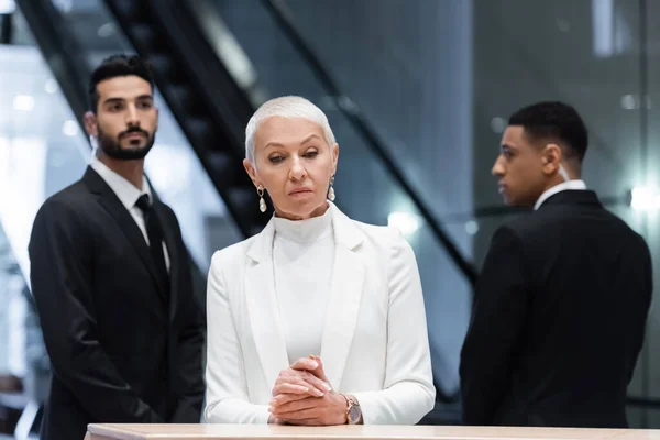 Senhora de negócios sênior elegante em pé na recepção do hotel perto de guarda-costas interracial borrada — Fotografia de Stock