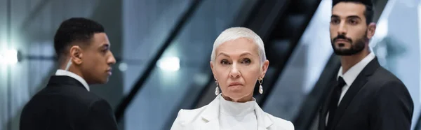 Senior businesswoman looking at camera near private interracial bodyguards in hotel, banner — Stock Photo