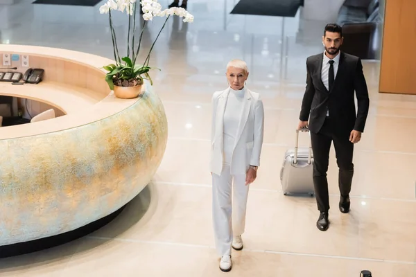Mixed race security man in black suit carrying suitcase near stylish senior businesswoman in hotel lobby — Stock Photo