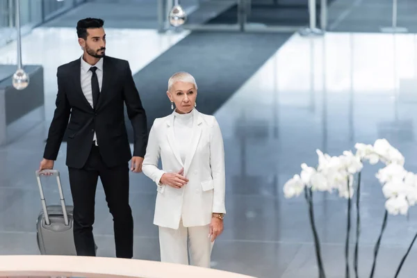 Mature business lady in white suit, and bi-racial bodyguard with suitcase near hotel reception — Stock Photo