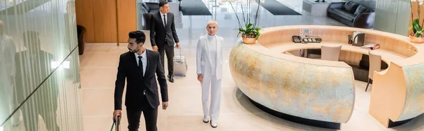 Высокий угол зрения стильной старшей предпринимательницы рядом с многонациональными частными телохранителями в вестибюле отеля, баннер — стоковое фото