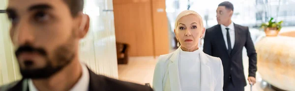 Selective focus of wealthy senior businesswoman near blurred multiethnic guards in hotel hall, banner — Stock Photo