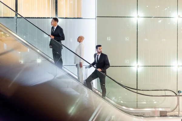 Femme d'affaires mature sur l'escalier roulant de l'hôtel près de gardes du corps interracial — Photo de stock