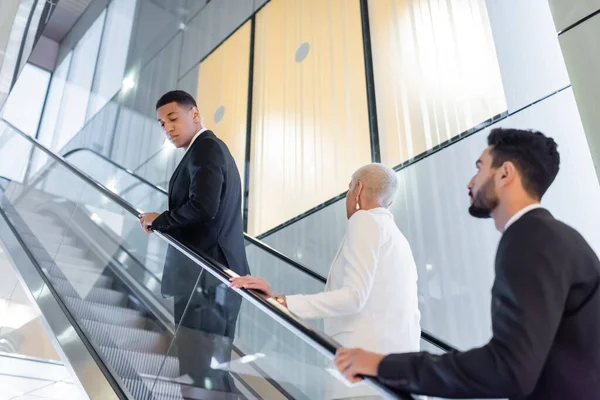 Interracial security men in formal wear escorting senior businesswoman on hotel escalator — Stock Photo