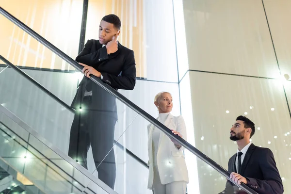 Femme d'affaires mature regardant loin près de gardes du corps multiethnique sur l'escalier roulant de l'hôtel — Photo de stock