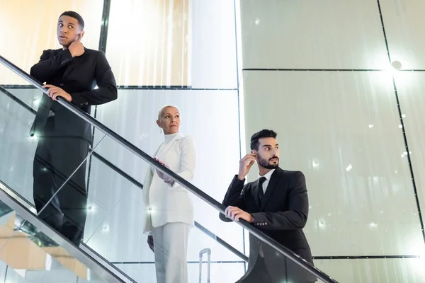 Interracial bodyguards adjusting earpiece near wealthy senior businesswoman on hotel escalator — Stock Photo