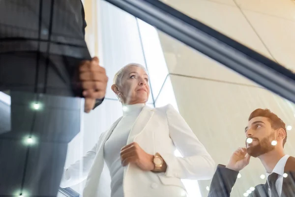 Vue à faible angle de la femme d'affaires senior élégant près de gardes du corps interracial sur l'escalier roulant de l'hôtel — Photo de stock