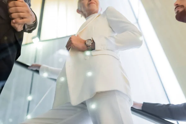 Low angle view of senior businesswoman in white clothes near interracial bodyguards on hotel escalator — Stock Photo