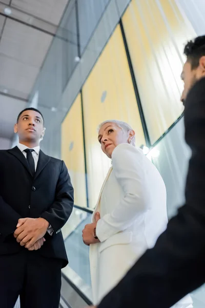 Vue à angle bas d'hommes de sécurité multiethniques escortant une femme d'affaires âgée sur un escalier roulant d'un hôtel — Photo de stock