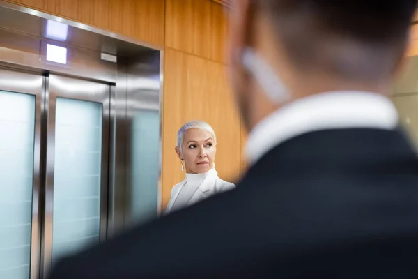 Senior business lady waiting for elevator in hotel near african american bodyguard on blurred foreground - foto de stock