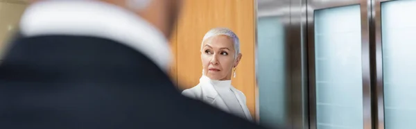 Reife Geschäftsfrau wartet auf Aufzug in Hotel in der Nähe von bi-rassischen Leibwächter auf verschwommenem Vordergrund, Banner — Stockfoto