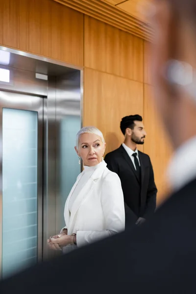 Hombre de seguridad superior esperando ascensor cerca de guardaespaldas bi-racial y hombre de seguridad afroamericano en primer plano borroso - foto de stock