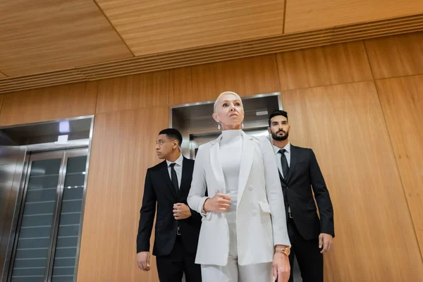 Low angle view of successful mature business lady near multiethnic bodyguards in hotel hall — Stock Photo