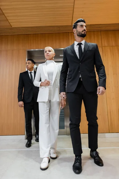 Full length view of stylish senior business lady walking near private interracial bodyguards in hotel hall — Stock Photo