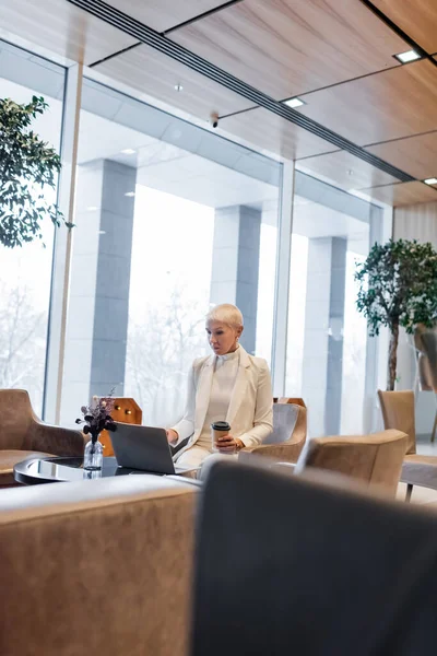 Dame d'affaires senior avec boisson à emporter en utilisant un ordinateur portable dans le salon de l'hôtel — Photo de stock