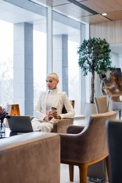 Старша бізнес-леді з паперовою чашкою, що сидить біля комп'ютера в готельному залі — стокове фото