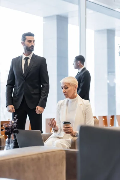 Mujer de negocios senior con café para ir apuntando a la computadora portátil cerca borrosa guardias multiculturales - foto de stock