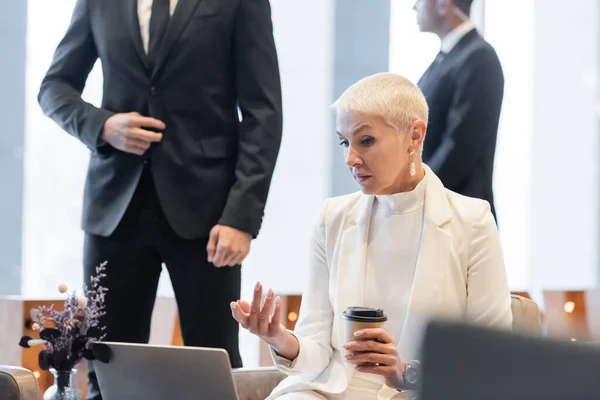 Mujer de negocios senior con taza de papel apuntando a la computadora portátil cerca borrosa guardaespaldas multiétnicos - foto de stock