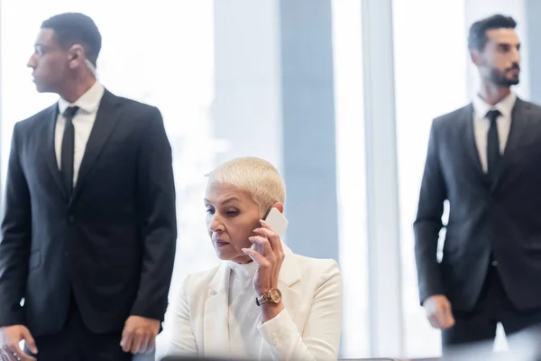 Des gardes du corps multiethniques flous protégeant une femme d'affaires âgée parlant sur un smartphone dans le hall de l'hôtel — Photo de stock