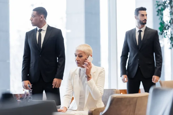 Mulher de negócios madura chamando no smartphone perto de laptop e guardas inter-raciais no hotel — Fotografia de Stock