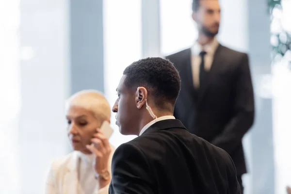 Guardaespaldas afroamericano cerca de mujer de negocios madura hablando en el teléfono inteligente en el hotel - foto de stock